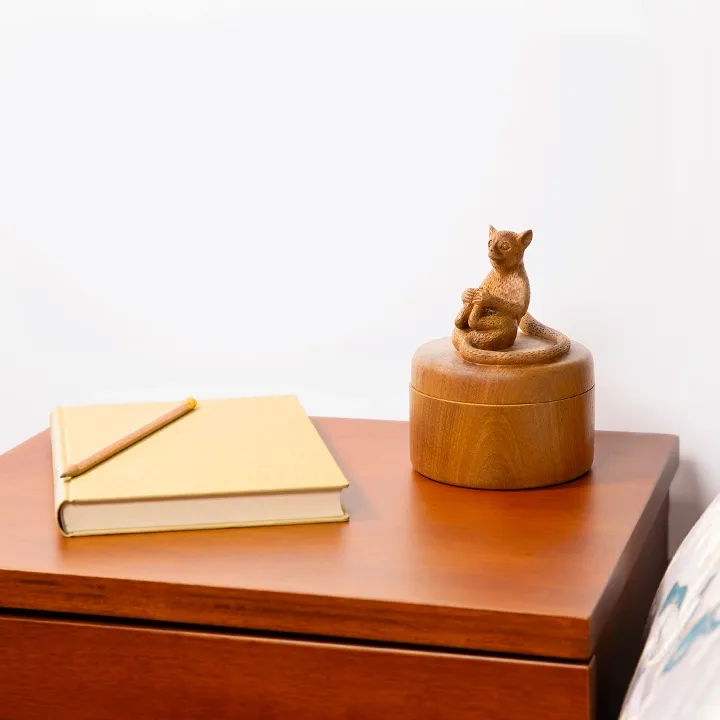 A decorative bedside item: a round, handcrafted wooden jewelry box adorned with a handle in the shape of a seated lemur.