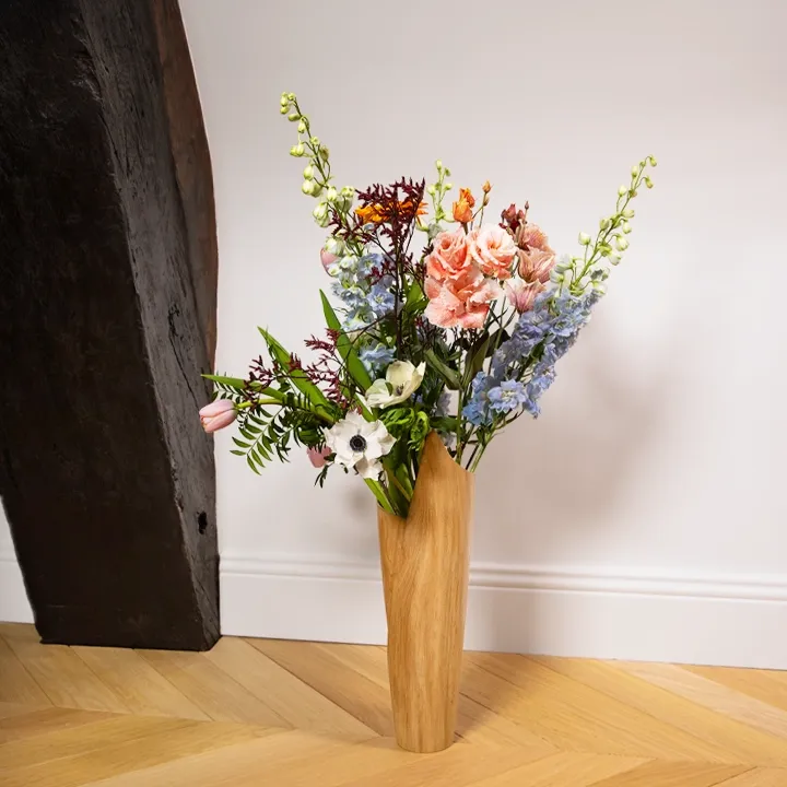 Tall, modern wooden vase with a unique folded design, holding a colorful floral arrangement on a wooden floor.